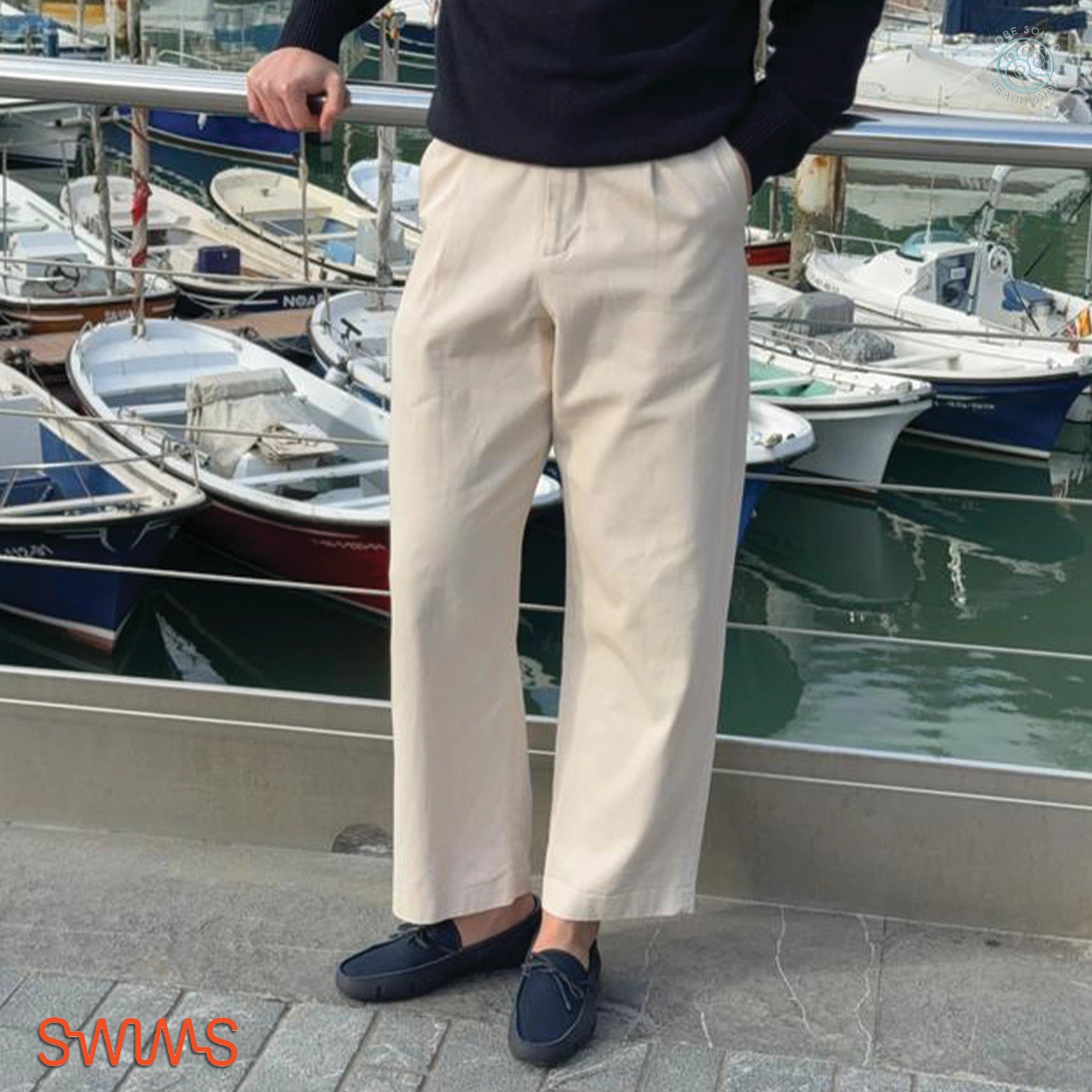 Person wearing beige pants and Swims Navy Loafers standing by a marina with boats in the background.
