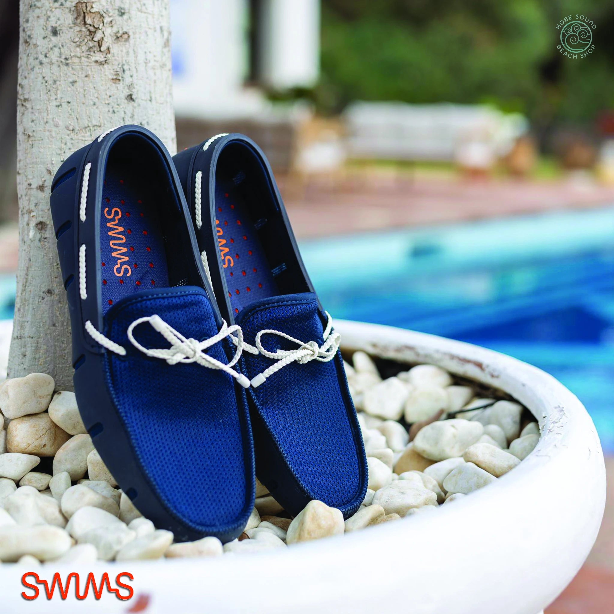 Blue Swims loafers with white laces on pebbles near a pool.