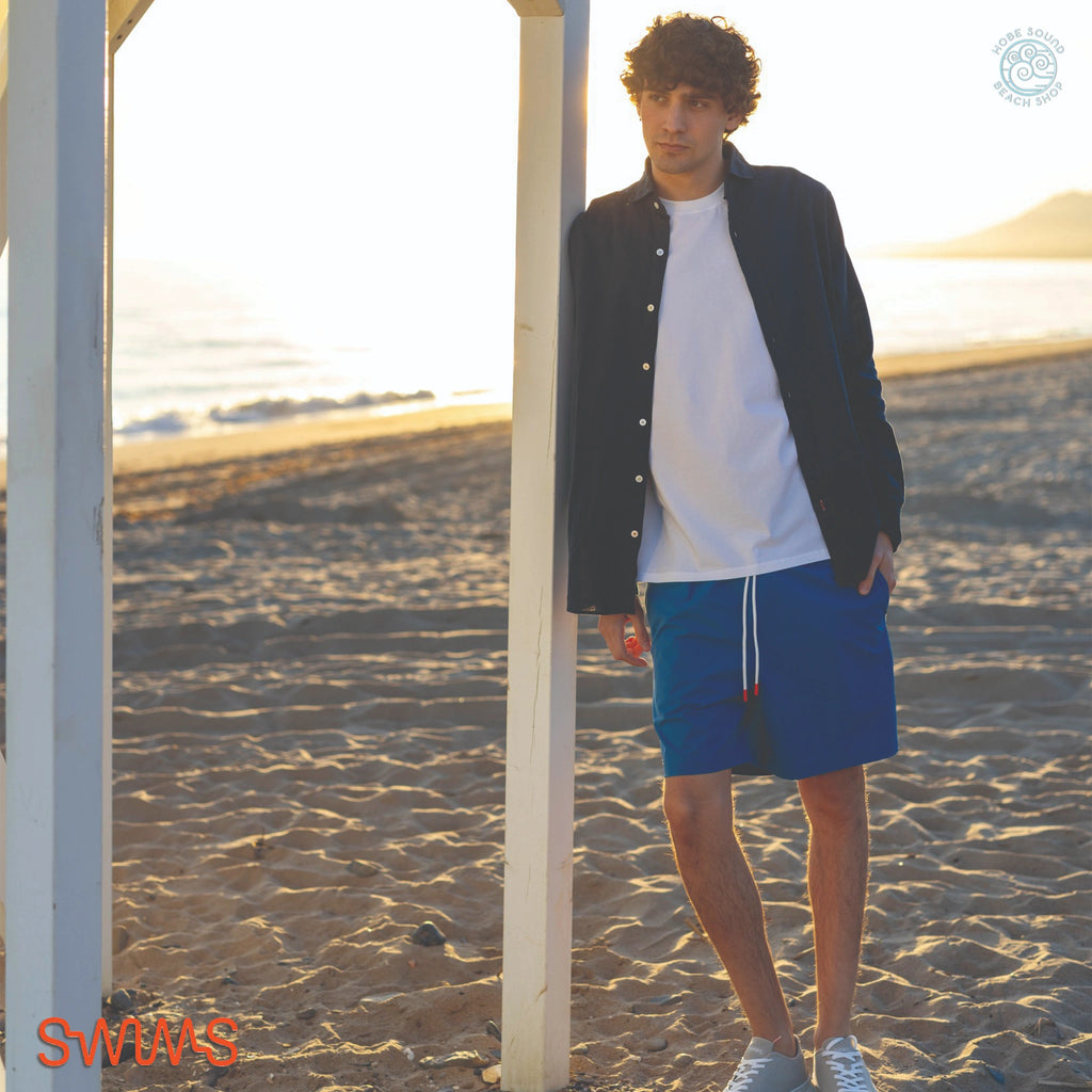 Man standing on a beach wearing a SWIMS Santorini Linen Shirt over a white t-shirt, and blue shorts.