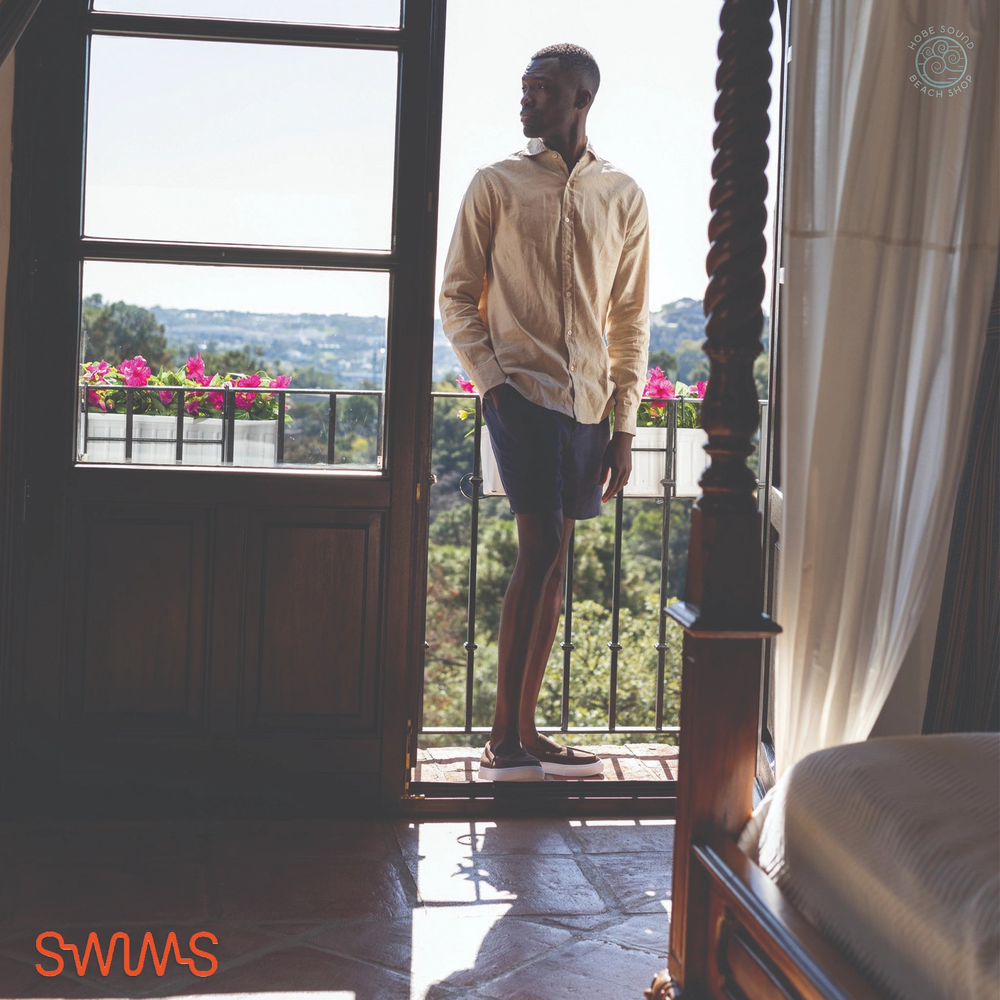 Man standing in a room with a large window overlooking a scenic view, wearing a beige shirt and SWIMS Samos Shorts.