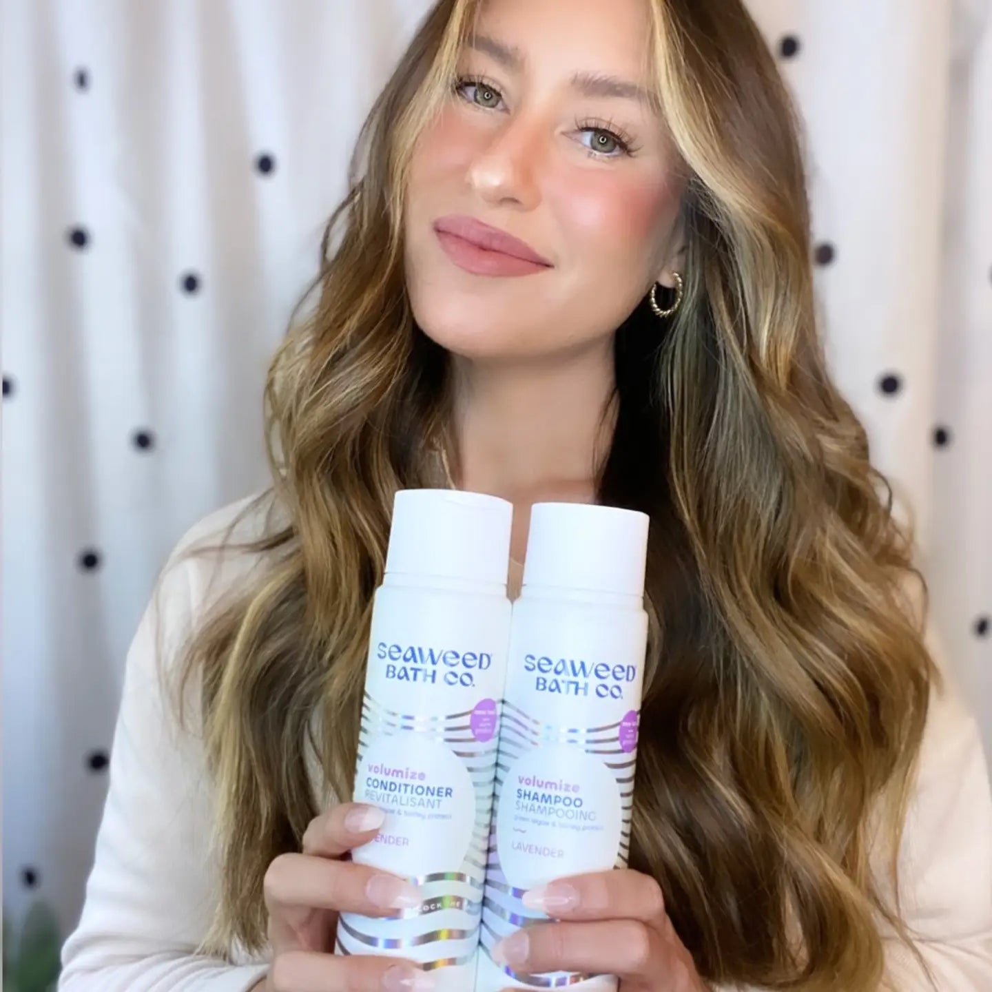 Woman holding two bottles of SeaWeed Bath Co. shampoo and conditioner against a polka dot curtain background.